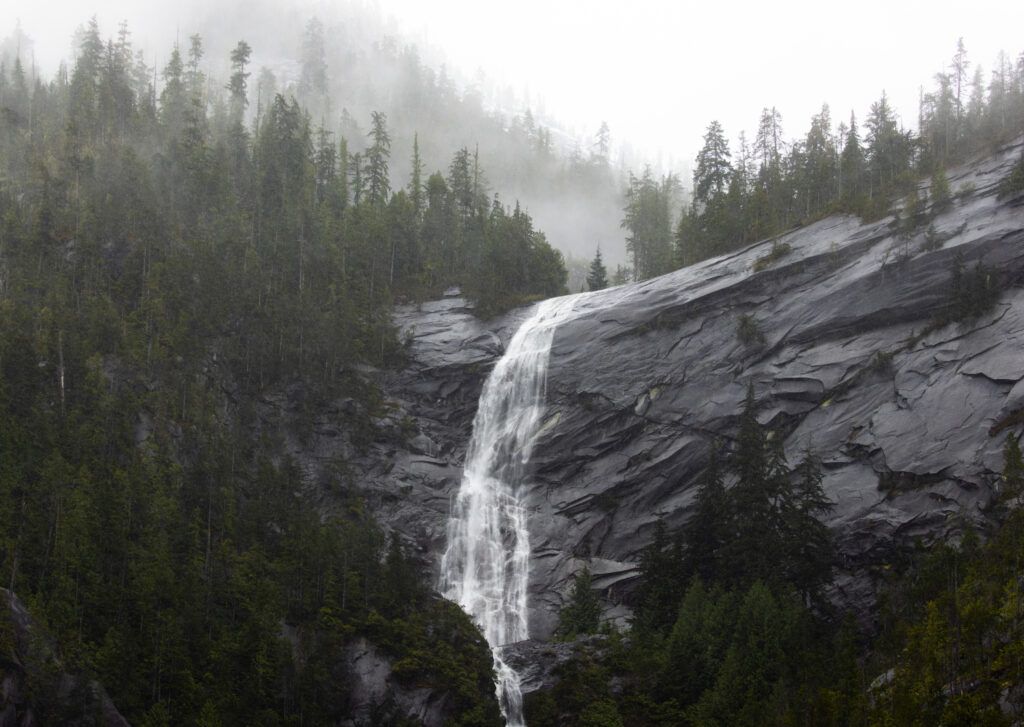 Waterfall following over a large rock face with thick forest on top. There is haze in the trees.