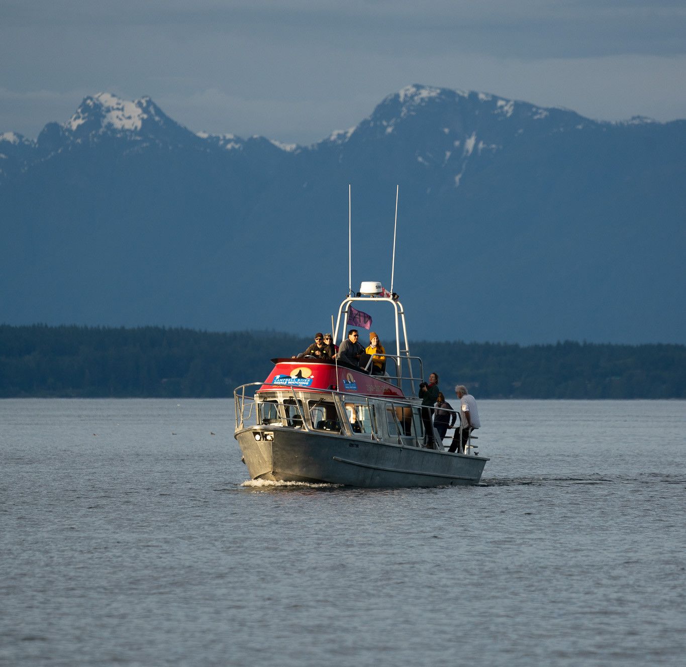 Covered Whale Watching Vessel out in the ocean. There are mountains in the foreground. There are guests on the the lower deck as well as the upper viewing deck.