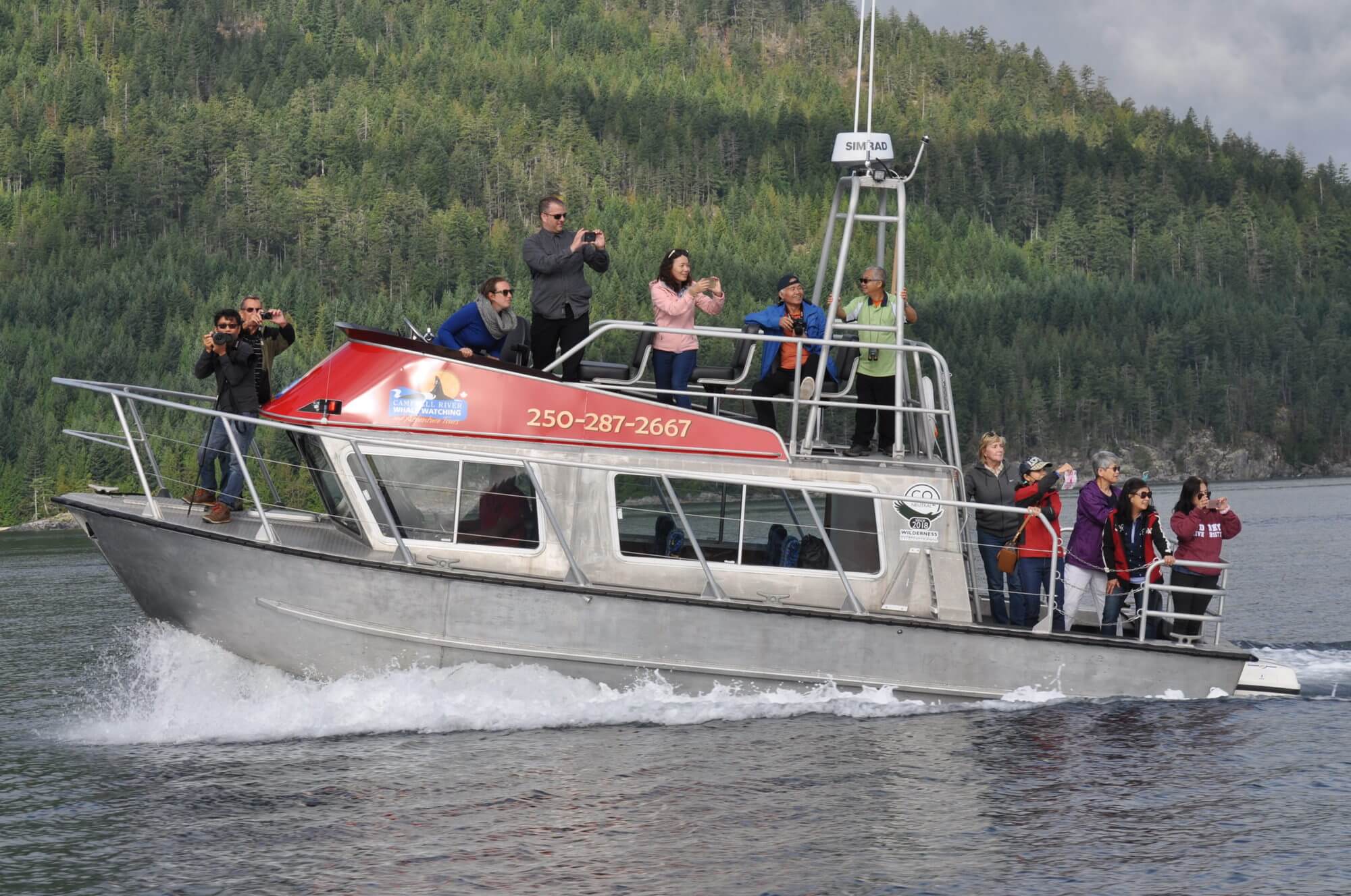 Image of a covered whale watching boat underway. There are two guests out on the lower and upper decks taking photos and looking at something not pictured.